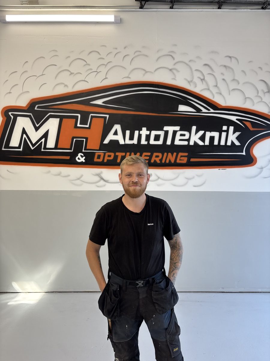 Marcus Hvitfeldt, ejer af MH Autoteknik Aps, foran værkstedets logo