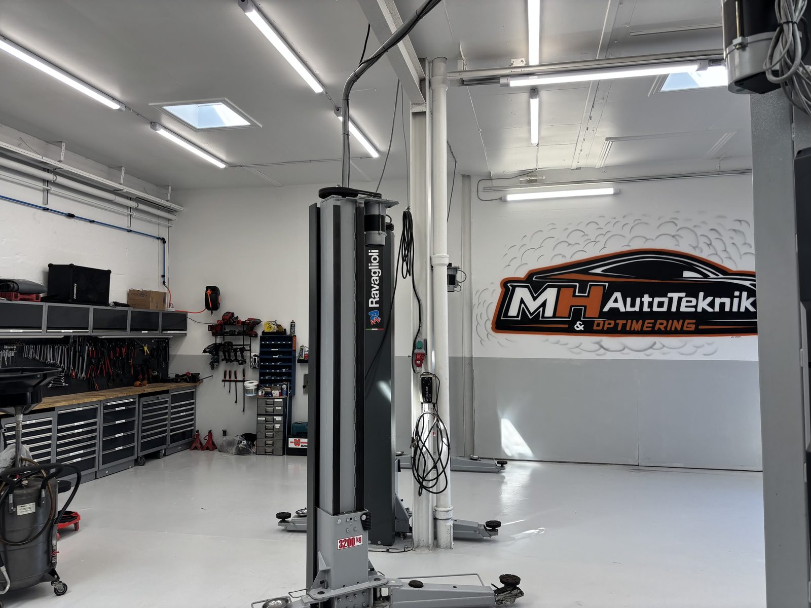 Værksted hos MH Autoteknik med lift og MH Autoteknik-logo på væggen